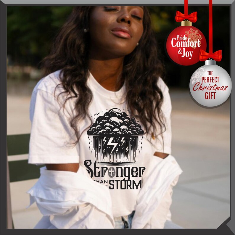 K&ouml;nnte beinhalten: Wei&szlig;es T-Shirt mit einem schwarz-wei&szlig;en Grafikdesign einer Gewitterwolke mit Blitzen und dem Text "Stronger Than Storm". Das Shirt hat einen Rundhalsausschnitt und kurze &Auml;rmel. Das Bild enth&auml;lt auch Weihnachtsschmuck.