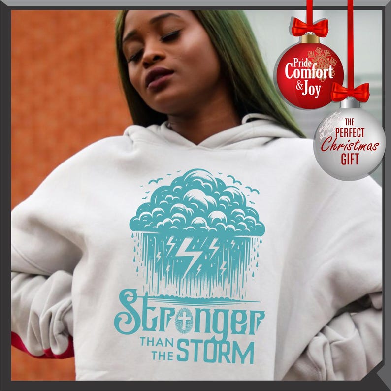 K&ouml;nnte beinhalten: Wei&szlig;er Kapuzenpullover mit einem t&uuml;rkisfarbenen Grafikdesign einer Gewitterwolke, Blitzen und dem Text "Stronger Than The Storm". Der Hoodie hat eine Kapuze mit Kordelzug und lange &Auml;rmel. Weihnachtsornamente sind ebenfalls zu sehen.