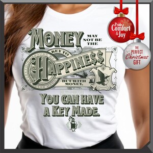 Pu&ograve; includere: T-shirt bianca con un design grafico con le parole "I soldi potrebbero non essere la chiave della felicit&agrave;, ma con i soldi, puoi farti fare una chiave". Il design include immagini di banconote e una piccola illustrazione di una persona che lavora in una fucina.