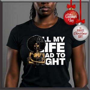 Może przedstawiać: Czarny t-shirt z białym napisem "ALL MY LIFE HAD TO FIGHT". Koszulka przedstawia grafikę kobiety z afro. Dwie bombki choinkowe wiszą w prawym g&oacute;rnym rogu, jedna czerwona i jedna biała.