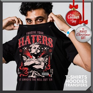 May include: Black t-shirt with the text "FORGIVE YOUR HATERS" in red, featuring an illustration of an angel with crossed arms. The shirt also has the text "IT ANNOYS THE HELL OUT 'EM". The image includes Christmas ornaments and text that says "T-SHIRTS HOODIES TRANSFERS".