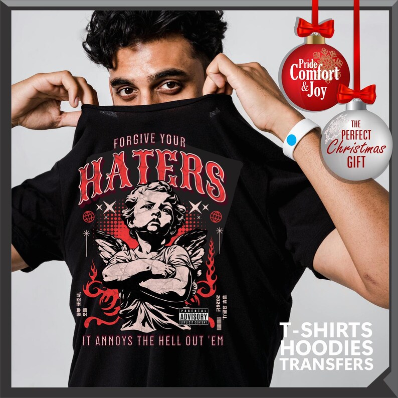 May include: Black t-shirt with the text "FORGIVE YOUR HATERS" in red, featuring an illustration of an angel with crossed arms. The shirt also has the text "IT ANNOYS THE HELL OUT 'EM". The image includes Christmas ornaments and text that says "T-SHIRTS HOODIES TRANSFERS".