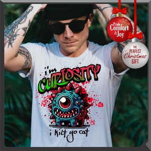 Może przedstawiać: Biała koszulka z kolorową postacią potwora z kresk&oacute;wki i napisem "I'm Curiosity i kilt yo cat". Potw&oacute;r jest niebieski, czerwony i zielony. Casualowy element odzieży z grafiką.