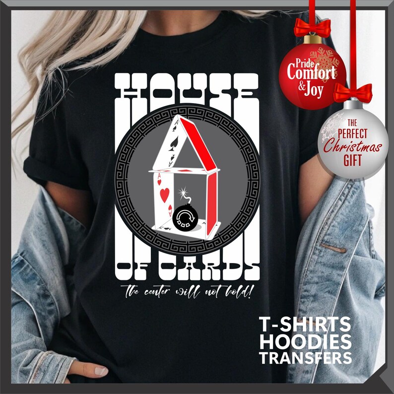 May include: Black t-shirt with a graphic of a house of cards, featuring playing card suits and a bomb. The text "HOUSE OF CARDS" is above the graphic, with "the center will not hold!" below. Additional text includes "T-SHIRTS HOODIES TRANSFERS".