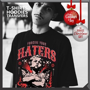 May include: Black t-shirt with the text "FORGIVE YOUR HATERS" in red, featuring an angel graphic. The image also includes text that says "T-SHIRTS HOODIES TRANSFERS". Two Christmas ornaments are also visible, one with the text "Pride Comfort & Joy" and the other with "THE PERFECT Christmas GIFT".