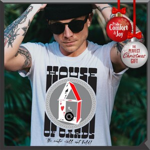 May include: White t-shirt with a graphic of a house made of playing cards, with the text "HOUSE OF CARDS" and "The center will not hold!". The design includes a red, white, and black color scheme. The shirt is a perfect Christmas gift.