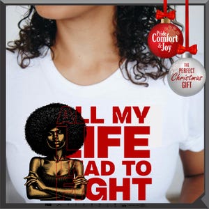 Może przedstawiać: Biała koszulka z grafiką kobiety z afro i czerwonym napisem "ALL MY LIFE HAD TO FIGHT". Dwie bombki choinkowe wiszą u g&oacute;ry po prawej stronie, jedna czerwona z napisem "Pride & Joy", a druga srebrna z napisem "THE PERFECT CHRISTMAS GIFT".