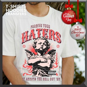 May include: White t-shirt with a graphic featuring an angel with crossed arms, the text "FORGIVE YOUR HATERS," and "IT ANNOYS THE HELL OUT 'EM." The design includes red accents and a parental advisory warning. The image also includes Christmas ornaments.