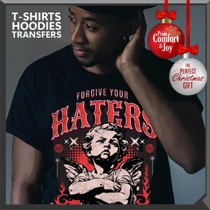 May include: Black t-shirt with the text "FORGIVE YOUR HATERS" in red, featuring an illustration of an angel. The image also includes the text "T-SHIRTS HOODIES TRANSFERS". Decorative Christmas ornaments with text are also visible.