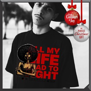 Może przedstawiać: Czarny t-shirt z grafiką kobiety z afro i napisem "ALL MY LIFE I HAD TO FIGHT" w kolorze czerwonym. Obraz zawiera r&oacute;wnież dwie bombki choinkowe z tekstem.