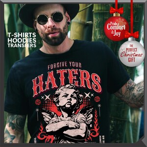 May include: Black t-shirt with the text "FORGIVE YOUR HATERS" in red, featuring an illustration of an angel. The image also includes text that says "T-SHIRTS HOODIES TRANSFERS". Two Christmas ornaments are also visible, one with the text "Pride Comfort & Joy" and the other with the text "THE PERFECT CHRISTMAS GIFT".