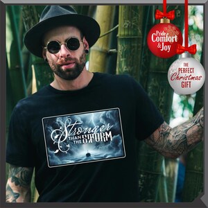 K&ouml;nnte beinhalten: Schwarzes T-Shirt mit einem rechteckigen Grafikdesign mit dem Text "Stronger Than The Storm" &uuml;ber einer st&uuml;rmischen Meereslandschaft mit einem Schiff. Das Bild enth&auml;lt auch zwei Weihnachtskugeln mit Text.