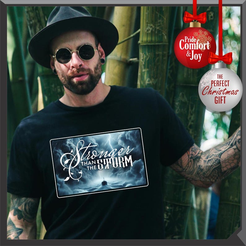 K&ouml;nnte beinhalten: Schwarzes T-Shirt mit einem rechteckigen Grafikdesign mit dem Text "Stronger Than The Storm" &uuml;ber einer st&uuml;rmischen Meereslandschaft mit einem Schiff. Das Bild enth&auml;lt auch zwei Weihnachtskugeln mit Text.