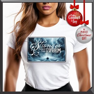 K&ouml;nnte beinhalten: Wei&szlig;es T-Shirt mit einem rechteckigen Aufdruck mit dem Text "Stronger Than The Storm" &uuml;ber einem Schiff, das durch st&uuml;rmische Gew&auml;sser segelt. Die Grafik ist von einem dunkelblauen Rand umgeben. Zwei Weihnachtskugeln h&auml;ngen oben.