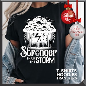 K&ouml;nnte beinhalten: Schwarzes T-Shirt mit einem wei&szlig;en Grafikdesign einer Gewitterwolke, Blitzen und dem Text "Stronger Than The Storm". Das Shirt hat auch den Text "T-SHIRTS HOODIES TRANSFERS" unten. Rote und wei&szlig;e Weihnachtskugeln im Hintergrund.