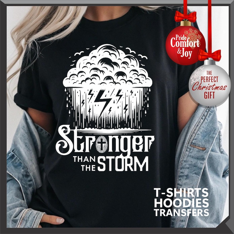 K&ouml;nnte beinhalten: Schwarzes T-Shirt mit einem wei&szlig;en Grafikdesign einer Gewitterwolke, Blitzen und dem Text "Stronger Than The Storm". Das Shirt hat auch den Text "T-SHIRTS HOODIES TRANSFERS" unten. Rote und wei&szlig;e Weihnachtskugeln im Hintergrund.