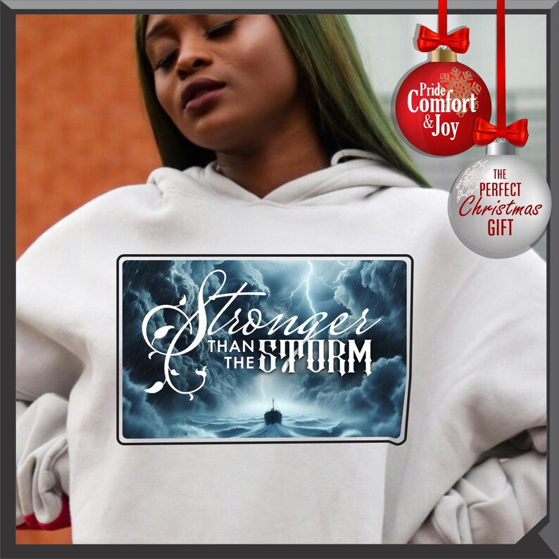 K&ouml;nnte beinhalten: Wei&szlig;er Kapuzenpullover mit dem Text "Stronger Than The Storm" auf der Vorderseite, &uuml;ber einer st&uuml;rmischen Ozeanszene mit einem Schiff. Der Kapuzenpullover hat eine Kapuze mit Kordelzug und lange &Auml;rmel. Weihnachtsbaumkugeln in Rot und Wei&szlig; sind im Hintergrund.