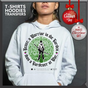 May include: White hoodie with a graphic featuring a figure holding a sword, surrounded by a green floral design. Text reads "Better, a Warrior in the Garden than a Gardener at War." The image includes text that says "T-SHIRTS HOODIES TRANSFERS".