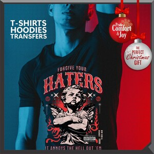 May include: Black t-shirt with the text "FORGIVE YOUR HATERS" and an illustration of an angel with crossed arms. The shirt also has the text "IT ANNOYS THE HELL OUT 'EM" and "ADVISORY". The image includes Christmas ornaments.