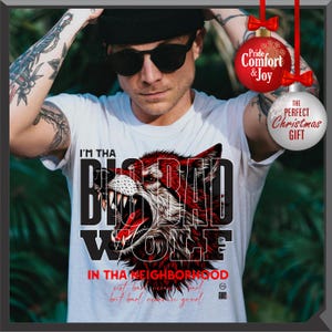 Op de afbeelding: Wit T-shirt met een afbeelding van een grommende rode wolf en de tekst "I'M THA BAD WOLF". Het shirt heeft ook de tekst "IN THA NEIGHBORHOOD" en "THE PERFECT CHRISTMAS GIFT". Een persoon draagt het shirt.