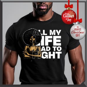 Może przedstawiać: Czarna koszulka z grafiką kobiety z afro i napisem "ALL MY LIFE HAD TO FIGHT" w kolorze białym i złotym. Dwie bombki choinkowe w tle.