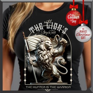 May include: Black t-shirt with a detailed lion illustration holding a flag. Text includes "THE LION'S Story is told" and "THE HUNTER IS THE WARRIOR". Features Christmas ornament graphics with "Pride Comfort & Joy" and "THE PERFECT Christmas GIFT".