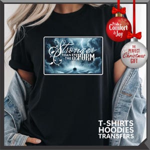 K&ouml;nnte beinhalten: Schwarzes T-Shirt mit einem rechteckigen Grafikmotiv mit dem Text "Stronger than the Storm" &uuml;ber einer st&uuml;rmischen Ozeanszene. Das Shirt hat auch den Text "T-SHIRTS HOODIES TRANSFERS" am unteren Rand. Rote und silberne Weihnachtsschmuck im Hintergrund.