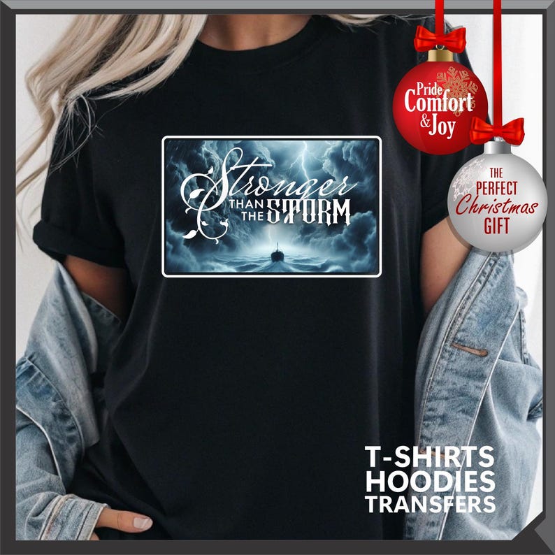 K&ouml;nnte beinhalten: Schwarzes T-Shirt mit einem rechteckigen Grafikmotiv mit dem Text "Stronger than the Storm" &uuml;ber einer st&uuml;rmischen Ozeanszene. Das Shirt hat auch den Text "T-SHIRTS HOODIES TRANSFERS" am unteren Rand. Rote und silberne Weihnachtsschmuck im Hintergrund.
