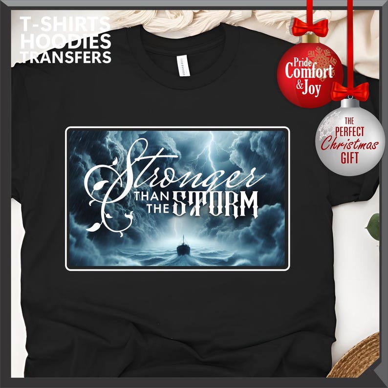 K&ouml;nnte beinhalten: Schwarzes T-Shirt mit einem rechteckigen Grafikmotiv mit der Aufschrift "Stronger Than The Storm" &uuml;ber einer st&uuml;rmischen Meereslandschaft mit einem Schiff. Das Shirt hat auch den Text "T-SHIRTS HOODIES TRANSFERS".