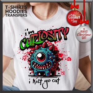 Może przedstawiać: Biała koszulka z kolorową grafiką potwora i napisem "I'm Curiosity i kict yo cat". Projekt przedstawia niebieskiego, czerwonego i zielonego potwora z jednym okiem i ostrymi zębami. Koszulka ma r&oacute;wnież napis "T-SHIRTS HOODIES TRANSFERS".