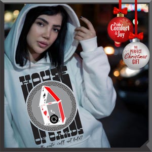 May include: White hoodie with a graphic of a house of cards, featuring playing card suits and the text "HOUSE OF CARDS". The text "The center will not hold!" is also printed on the hoodie. Red and white Christmas ornaments hang in the background.