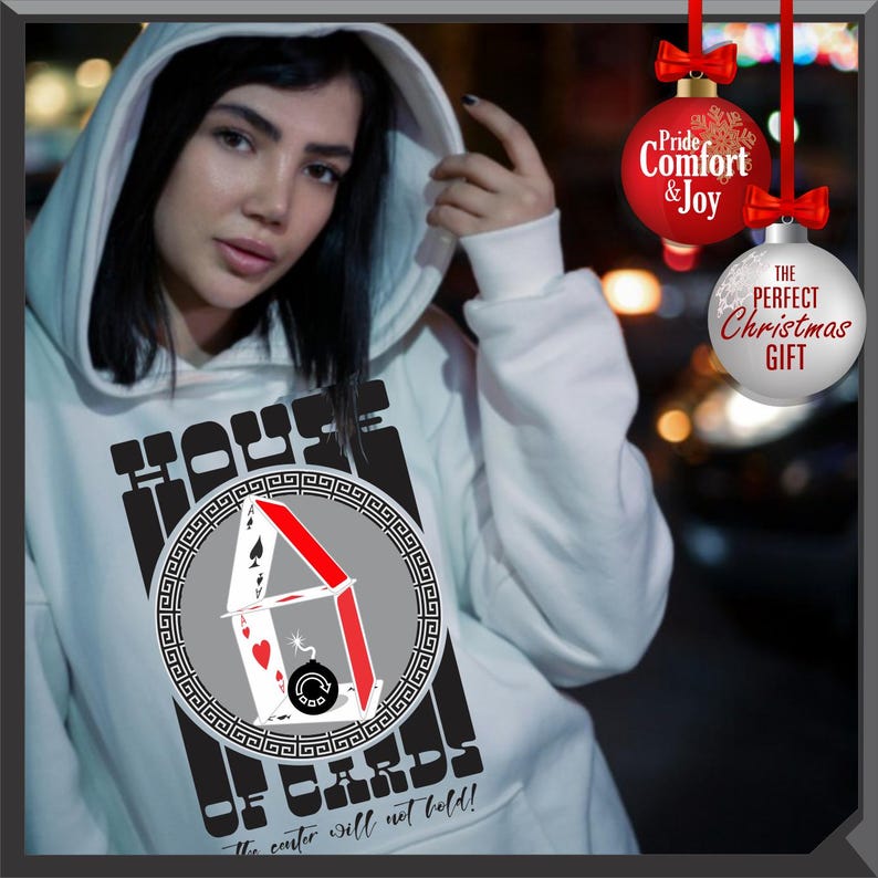 May include: White hoodie with a graphic of a house of cards, featuring playing card suits and the text "HOUSE OF CARDS". The text "The center will not hold!" is also printed on the hoodie. Red and white Christmas ornaments hang in the background.