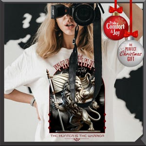 May include: White t-shirt with a lion graphic holding a flag, and the text "The Hunter is the Warrior" and "The Truth is Told". A person is wearing the shirt and holding a camera. Christmas decorations are visible in the background.