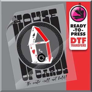 May include: A clear DTF transfer sheet featuring a graphic of a house of cards with a bomb. The text "HOUSE OF CARDS" and "The center will not hold!" are displayed. A red and white label reads "READY-TO-PRESS DTF TRANSFERS".