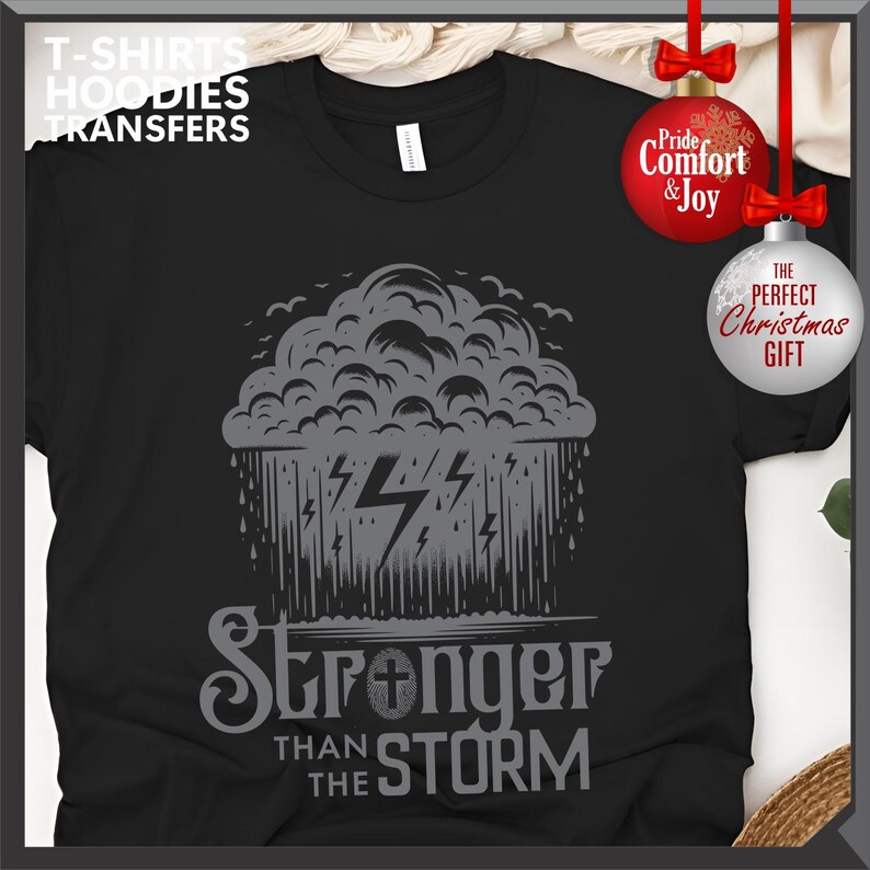 K&ouml;nnte beinhalten: Schwarzes T-Shirt mit einem grauen Design einer Gewitterwolke, Blitzen und dem Text "Stronger Than The Storm". Das Shirt hat auch den Text "T-SHIRTS HOODIES TRANSFERS". Das Bild enth&auml;lt Weihnachtsschmuck.