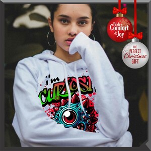 Może przedstawiać: Biała bluza z kapturem z grafiką z napisem "I'm Curious" w kolorze zielonym i czarnym oraz kolorowym wzorem oka potwora. Bluza z kapturem ma kaptur ze sznurkiem i długie rękawy. Obraz zawiera r&oacute;wnież ozdoby świąteczne.