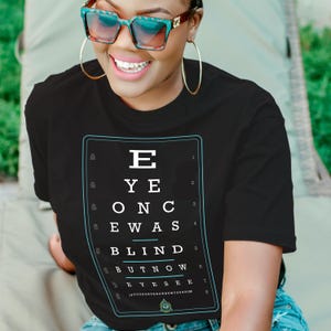 Puede incluir: Camiseta negra con un dise&ntilde;o de tabla optom&eacute;trica blanca. La tabla dice "EYE ONCE WAS BLIND BUT NOW EYESEE" en un marco rectangular. La persona de la imagen lleva gafas de sol y aros dorados.