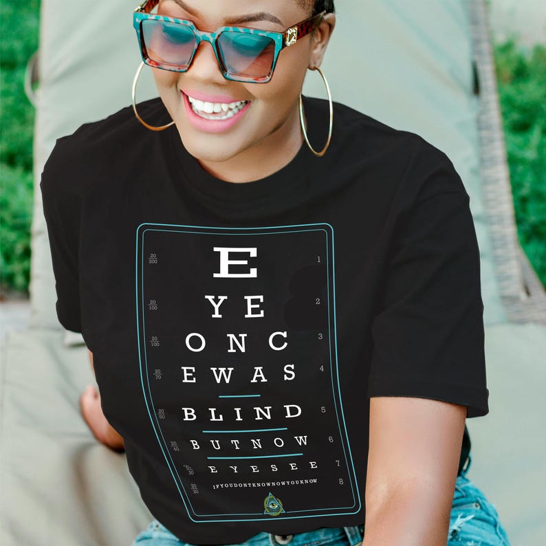 Puede incluir: Camiseta negra con un dise&ntilde;o de tabla optom&eacute;trica blanca. La tabla dice "EYE ONCE WAS BLIND BUT NOW EYESEE" en un marco rectangular. La persona de la imagen lleva gafas de sol y aros dorados.