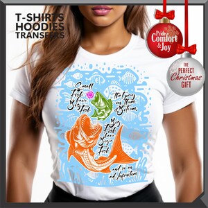 Pu&ograve; includere: T-shirt bianca con una grafica di pesce colorata e il testo "Small Fish have Big Fish" e "Big Fish have Bigger Fish". Il design include illustrazioni di pesci arancioni, verdi e blu. La maglietta ha anche il testo "The prey on them & bite 'em" e "and so on ad infinitum."