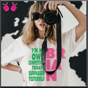 Puede incluir: Camiseta blanca con el texto "I'M MY OWN COMPETITION TODAY SURPASSED YESTERSELF" en verde y rosa. Una correa de c&aacute;mara negra cuelga frente a la camiseta. El fondo es un patr&oacute;n en blanco y negro.
