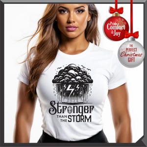 K&ouml;nnte beinhalten: Wei&szlig;es T-Shirt mit einer schwarz-wei&szlig;en Grafik einer Gewitterwolke, Blitzen und dem Text "Stronger Than The Storm". Das Shirt hat kurze &Auml;rmel und einen Rundhalsausschnitt.