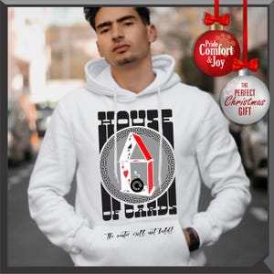 May include: White hoodie with a graphic of a house of cards, featuring playing cards and a bomb. The text reads "HOUSE OF CARDS" and "The center will not hold!". Includes a Christmas ornament that says "Pride, Comfort & Joy" and "The Perfect Christmas Gift".
