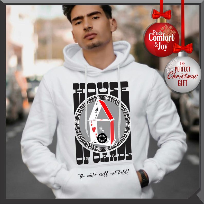 May include: White hoodie with a graphic of a house of cards, featuring playing cards and a bomb. The text reads "HOUSE OF CARDS" and "The center will not hold!". Includes a Christmas ornament that says "Pride, Comfort & Joy" and "The Perfect Christmas Gift".