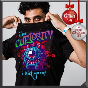 Może przedstawiać: Czarny t-shirt z kolorową grafiką potwora i napisem "I'm Curiosity i kilt yo cat". Projekt przedstawia jednoocznego stwora z niebieskimi, fioletowymi i czerwonymi detalami. Koszulka jest trzymana przez osobę.