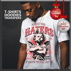 May include: White t-shirt with a graphic of an angel and the text "FORGIVE YOUR HATERS". The shirt also has the text "IT ANNOYS THE HELL OUT 'EM". The image includes text that says "T-SHIRTS, HOODIES, TRANSFERS".