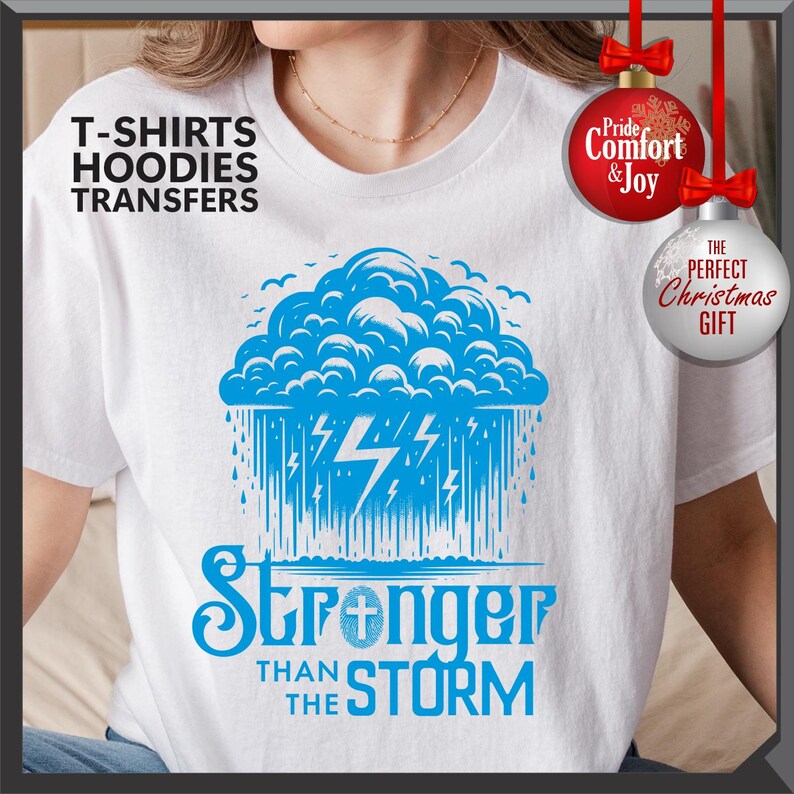 K&ouml;nnte beinhalten: Wei&szlig;es T-Shirt mit einem blauen Grafikdesign eines Sturms, Blitzen und Regen. Darunter steht der Text "Stronger Than The Storm". Das Shirt hat auch den Text "T-SHIRTS HOODIES TRANSFERS".