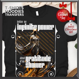 Może przedstawiać: Czarny t-shirt z grafiką postaci w powł&oacute;czystej sukni, złotymi akcentami oraz napisami "infinite power" i "gratitude shower". Koszulka ma r&oacute;wnież napis "T-SHIRTS HOODIES TRANSFERS". W tle ozdoby świąteczne.