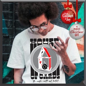 May include: White t-shirt with a graphic of a house of cards design. The design includes playing cards, a bomb, and the text "HOUSE OF CARDS" and "The center will not hold!". Two Christmas ornaments are in the background.