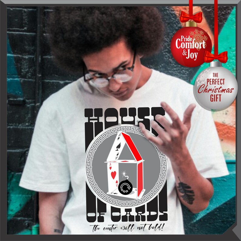 May include: White t-shirt with a graphic of a house of cards design. The design includes playing cards, a bomb, and the text "HOUSE OF CARDS" and "The center will not hold!". Two Christmas ornaments are in the background.