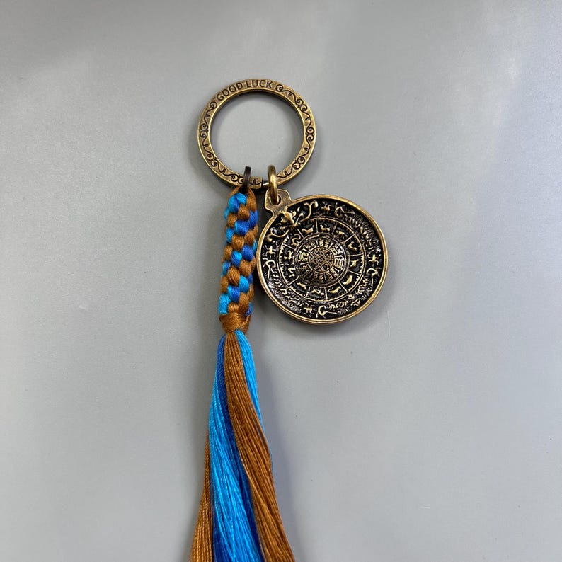Puede incluir: Un llavero de lat&oacute;n con un borla azul y marr&oacute;n. El llavero presenta un dije redondo con las palabras "Good Luck" y un dise&ntilde;o circular con s&iacute;mbolos.